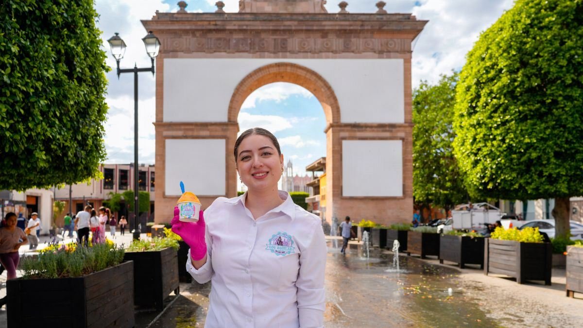 Una joven sonríe y sostiene un vaso de nieve artesanal colorida frente al histórico Arco de la Calzada durante la Feria de la Nieve León en León, Guanajuato.
