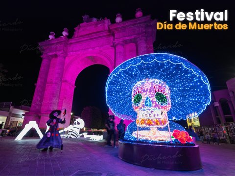 Calavera monumental luminosa con sombrero azul junto al Arco de la Calzada iluminado en rosa, en el Festival Día de Muertos León 2026